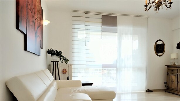 Un salon moderne avec un canapé blanc, une lampe sur pied, une plante en pot et une vue dégagée sur l’extérieur grâce à une grande fenêtre.