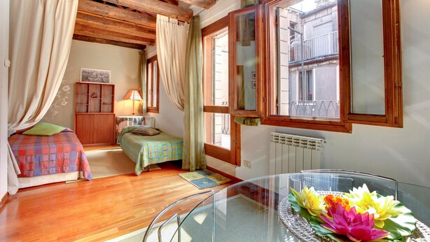 A bedroom with wooden beams, a bed with a colorful bedspread, a small table with a glass top, and a window with a view of a building. - Grimaldi Apartments - Ca Orlina (Venice)