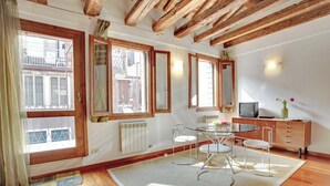A room with wooden beams, large windows, a dining table with chairs, a radiator, and a television on a cabinet. - Grimaldi Apartments - Ca Orlina (Venice)