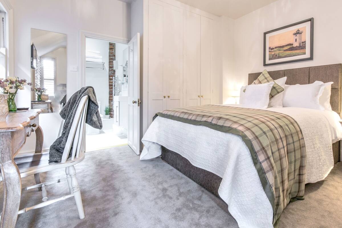 A bedroom with a bed, a nightstand, a lamp, a painting, and a door leading to a bathroom.