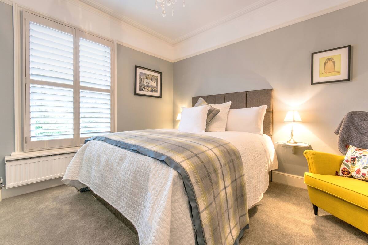 A bedroom with a bed, a yellow armchair, a nightstand with a lamp, and two framed pictures on the wall.