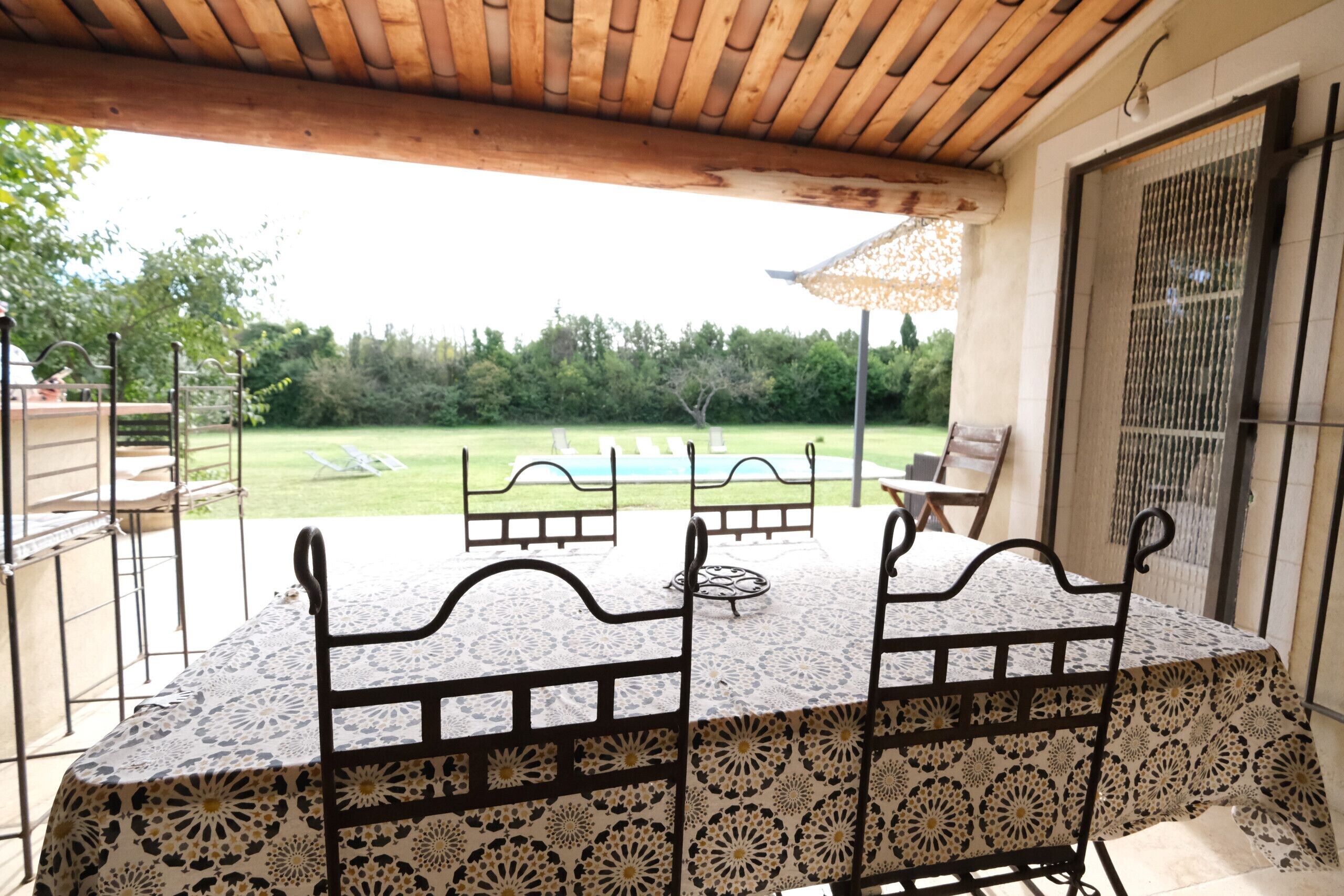 An outdoor patio with a table and chairs, a view of a swimming pool, and a covered area with a pergola.