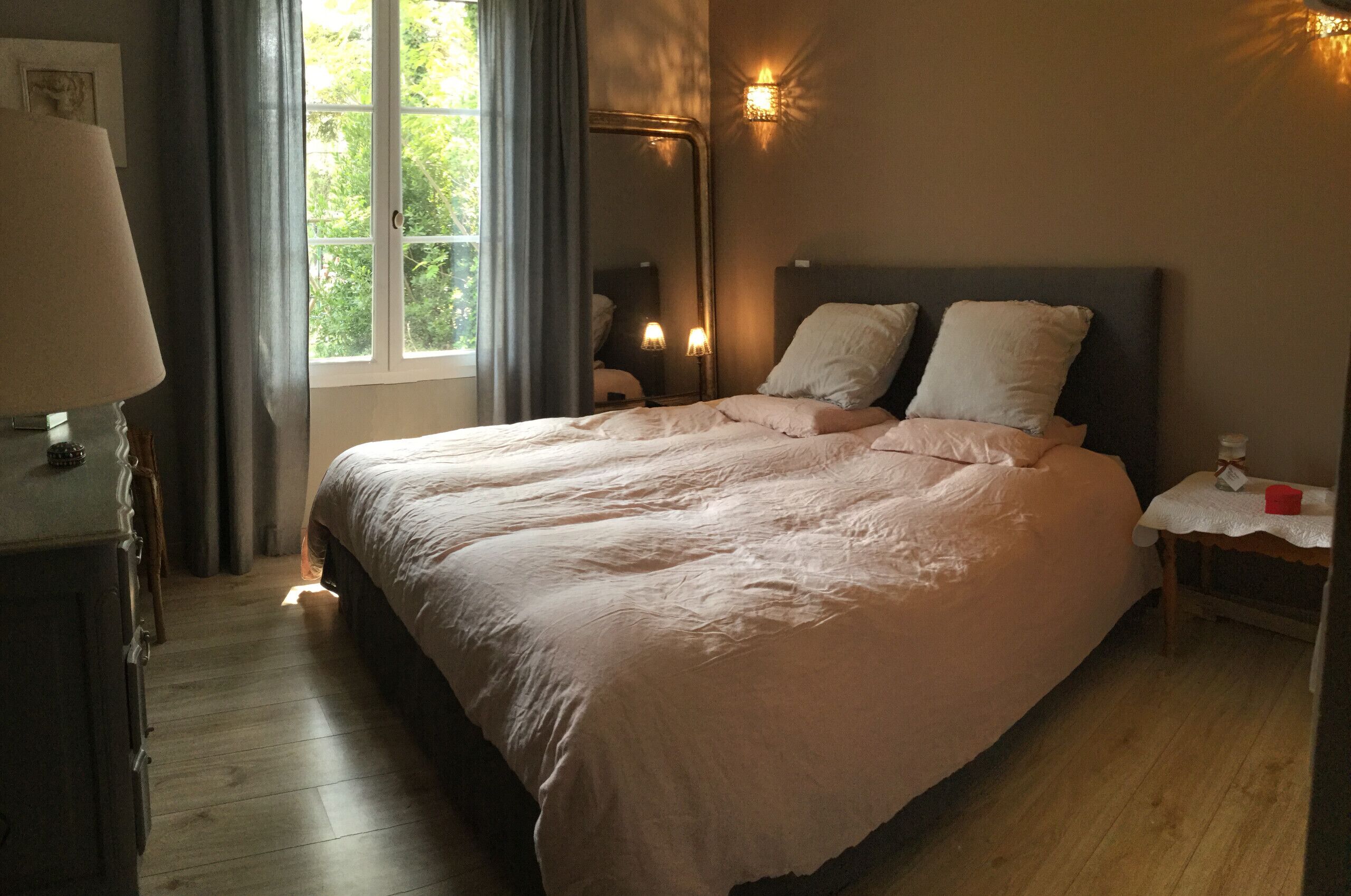A bedroom with a large bed, a nightstand, a lamp, a mirror, and a window with curtains.