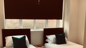 A hotel room with two single beds, each with white bedding and teal pillows. There is a window with blinds, a nightstand with a remote control, and a ceiling light. - Eazzzy rooms Corby (Corby)