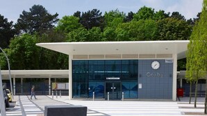 A modern building with a glass facade, a clock, and the name 'Corby' on the wall. There are benches and a pedestrian crossing in front. - Eazzzy rooms Corby (Corby)