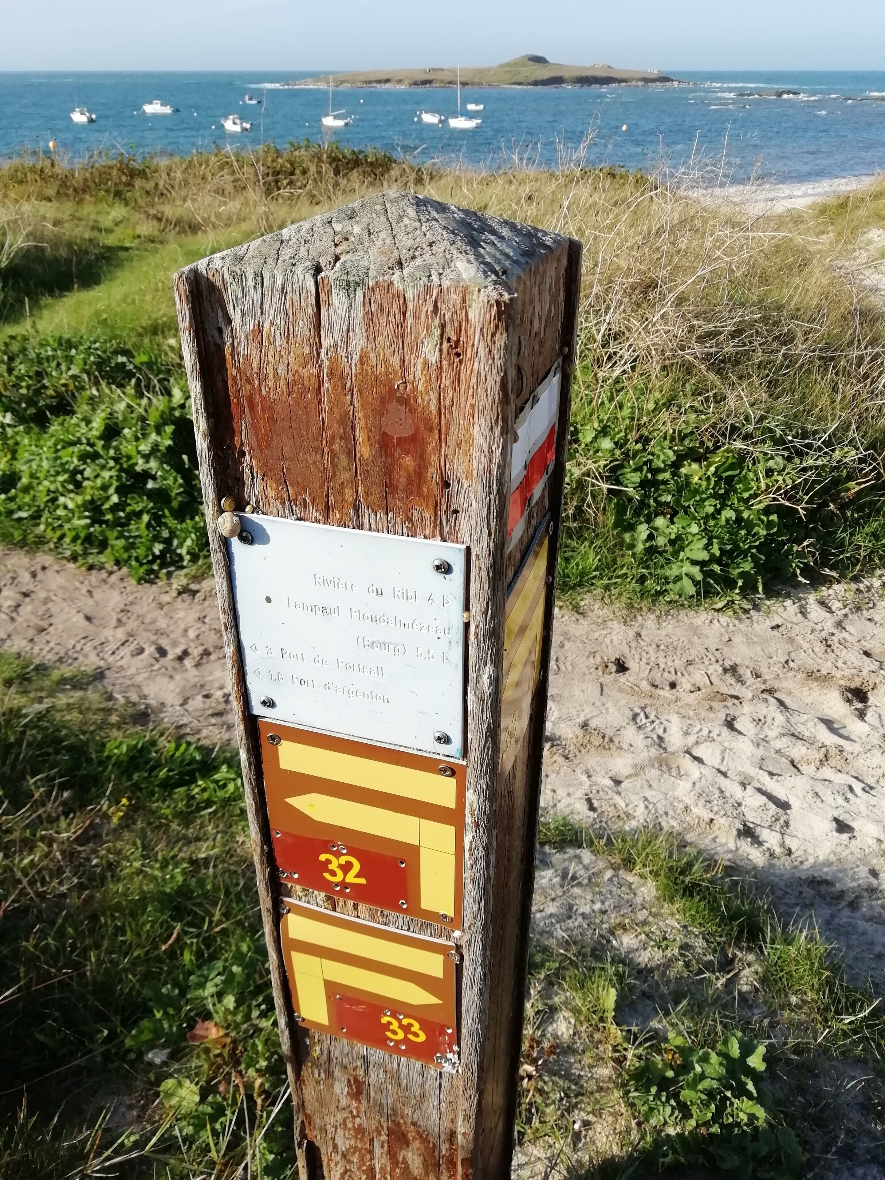 Een verweerde houten paal met richtingaanwijzingen en een bord met tekst in het Frans, nabij een strand met boten op de achtergrond.