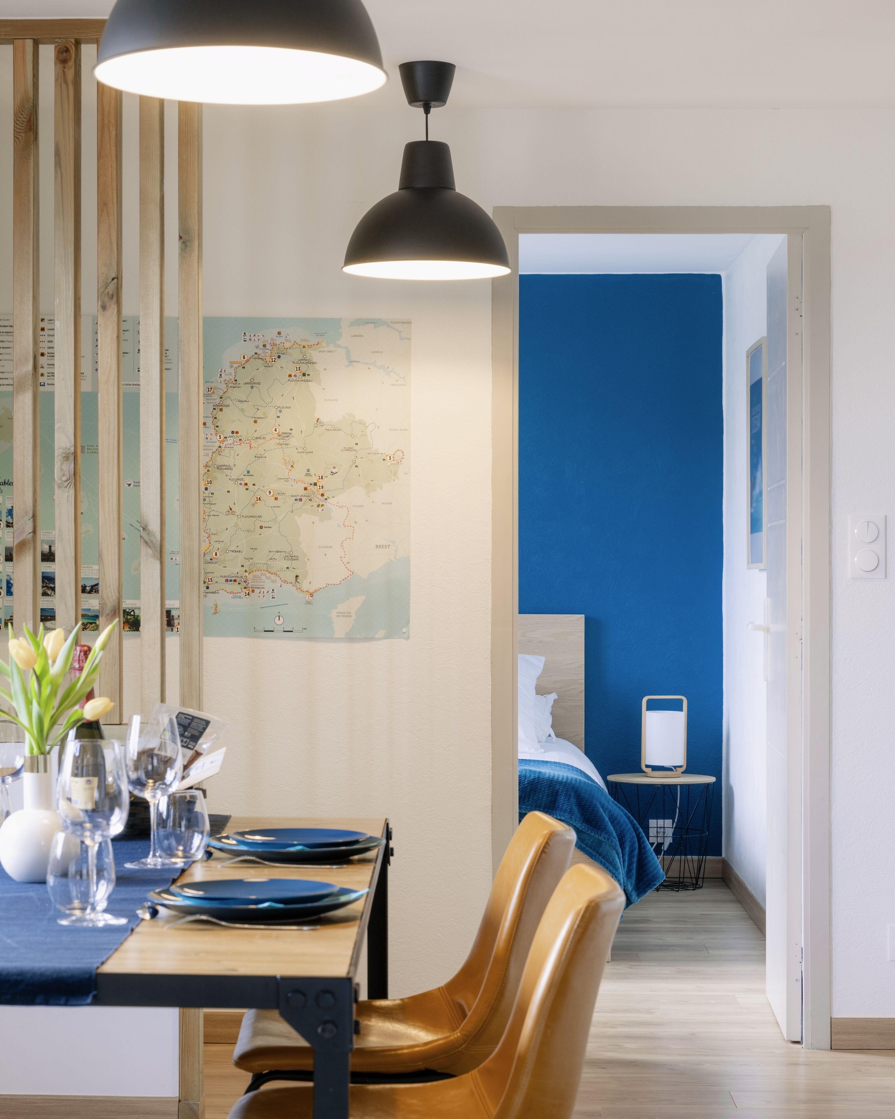 A hotel room with a wooden dining table, chairs, a map on the wall, a bed with blue bedding, and a desk with a computer.