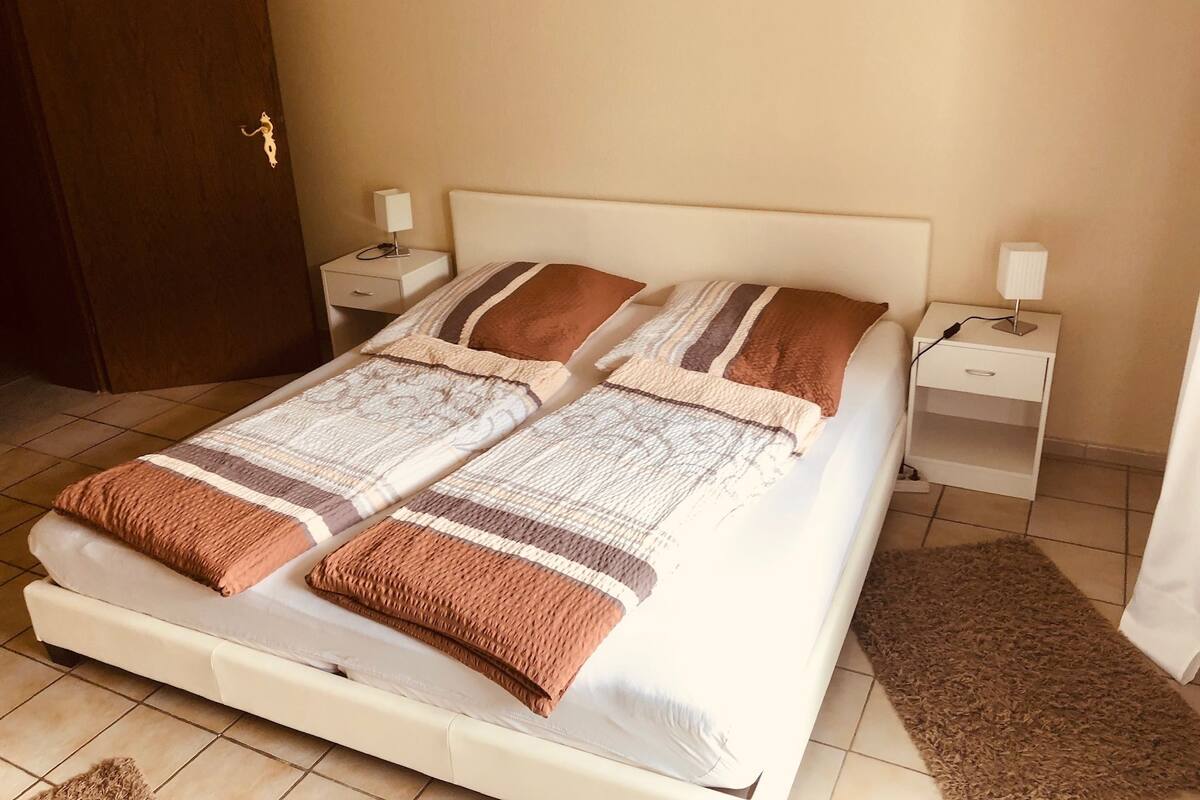 Un lit double avec une couvre-lit à motifs et deux oreillers, de chaque côté duquel se trouvent des tables de chevet avec des lampes, dans une pièce dont le sol est carrelé et qui possède une porte en bois.