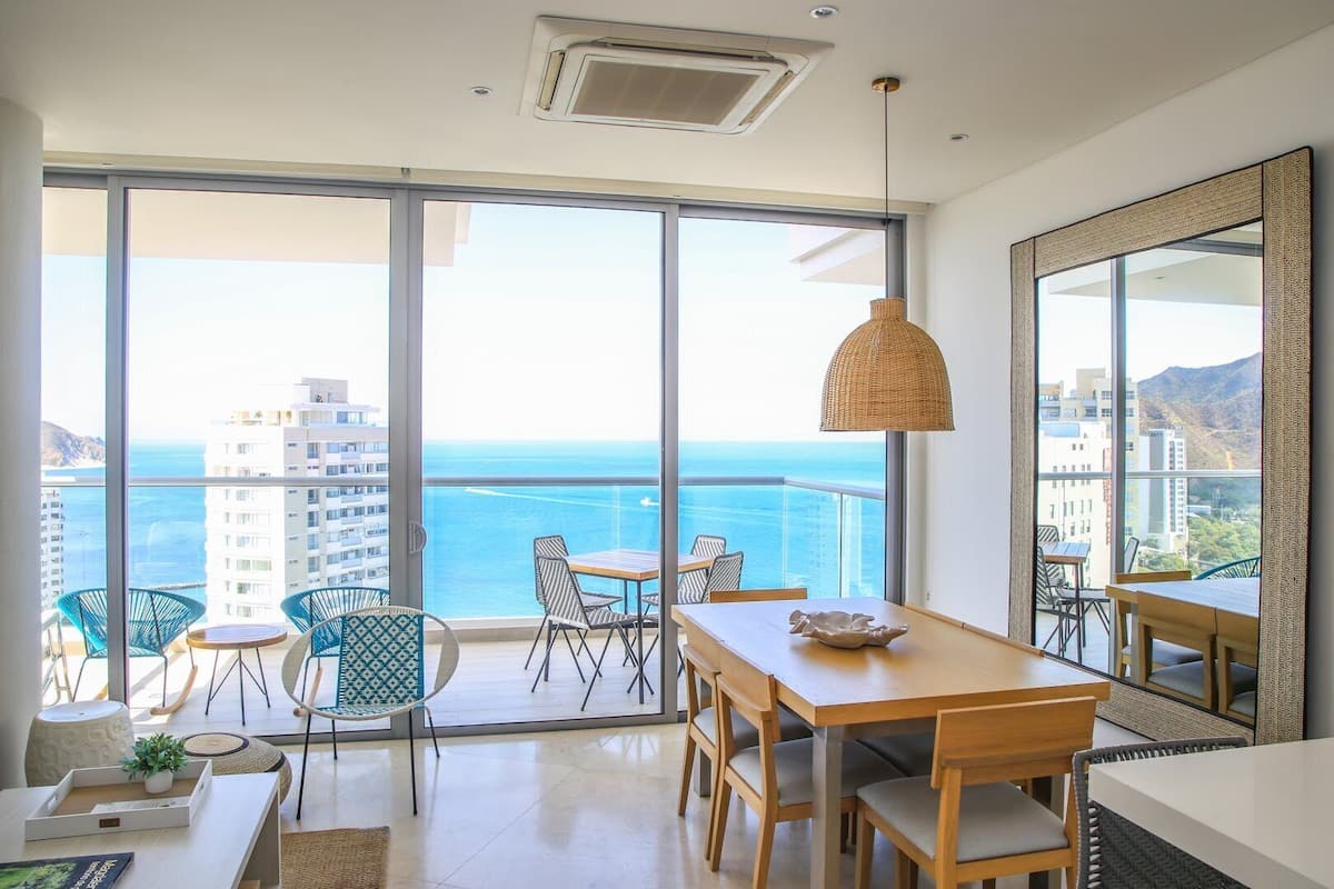 A spacious balcony with a view of the ocean and buildings, furnished with a table and chairs, and a hanging lamp.