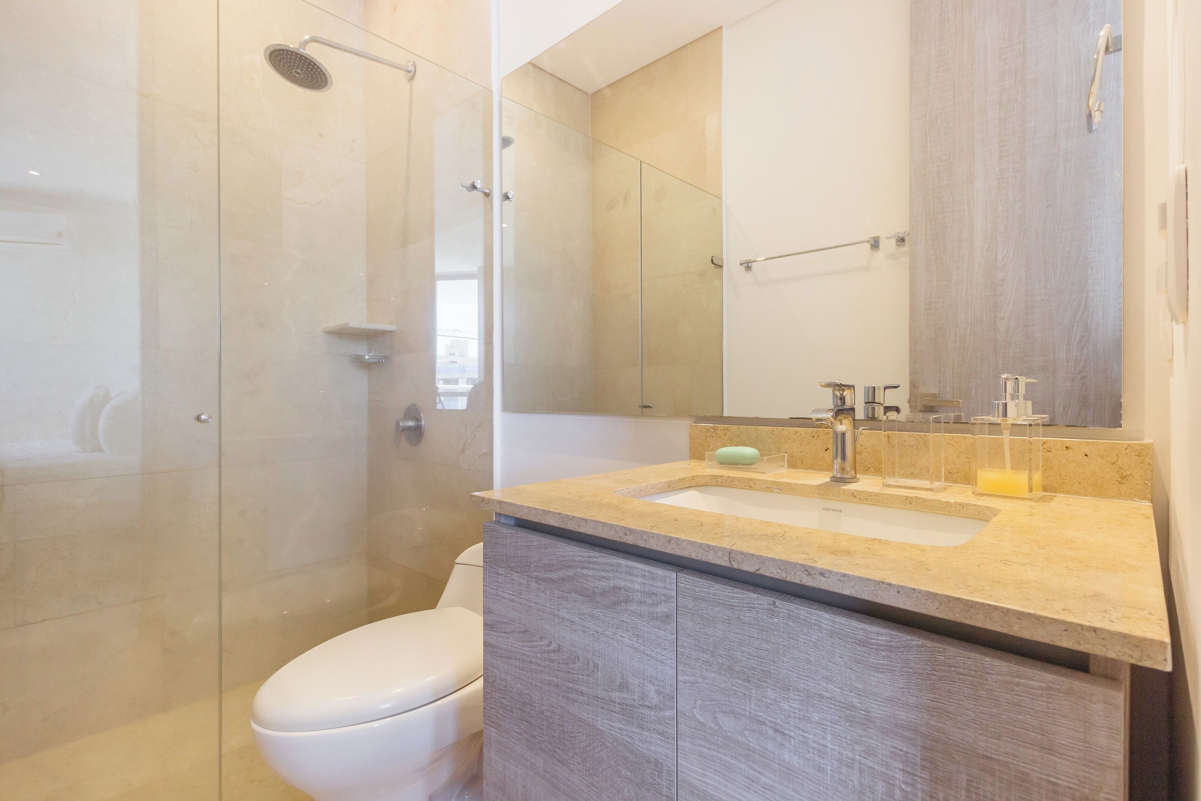 Un baño moderno con cabina de ducha de vidrio, inodoro blanco, encimera color beige con lavamanos y un espejo.