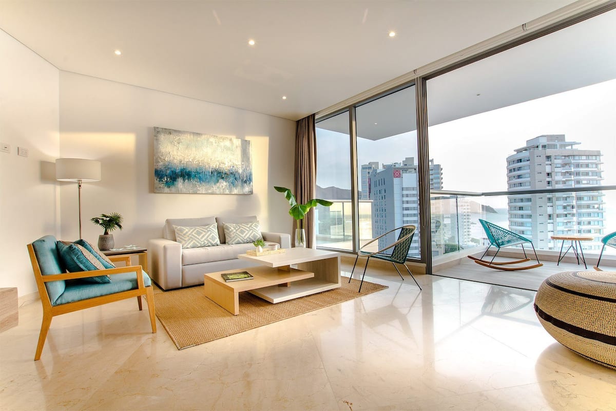 A modern living room with a sofa, coffee table, and chairs, large windows with a city view, and a painting on the wall.