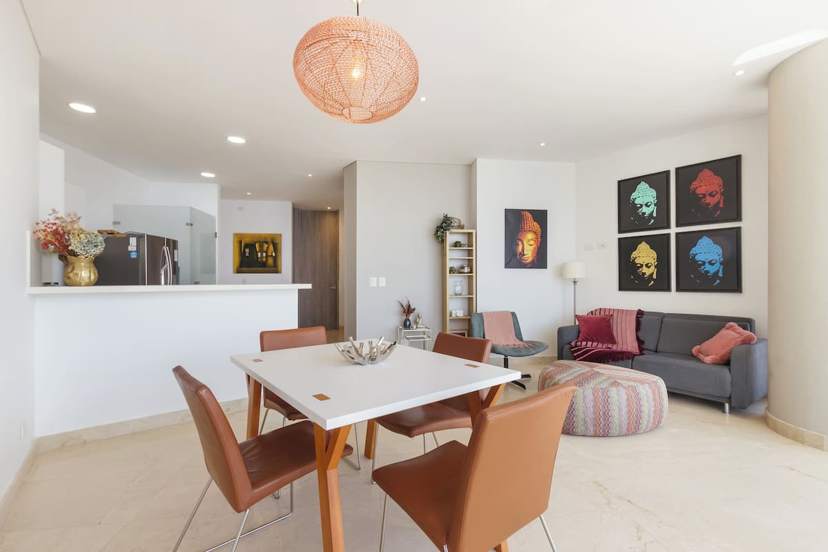 A modern living room with a dining area, featuring a white table, brown chairs, a grey sofa, and colorful artwork on the wall.