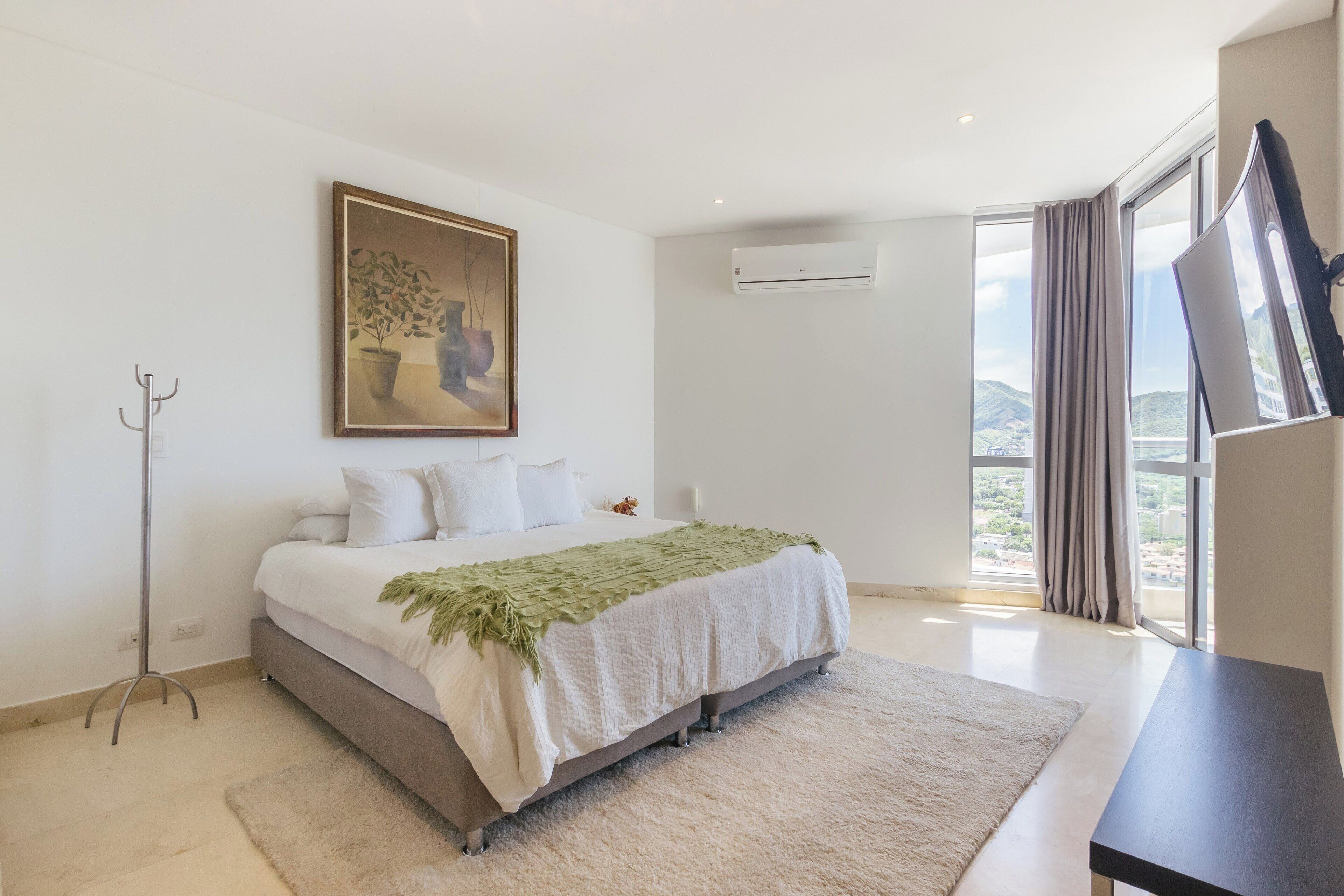 A bedroom with a bed, a painting, an air conditioning unit, a window with curtains, and a view of the outdoors.