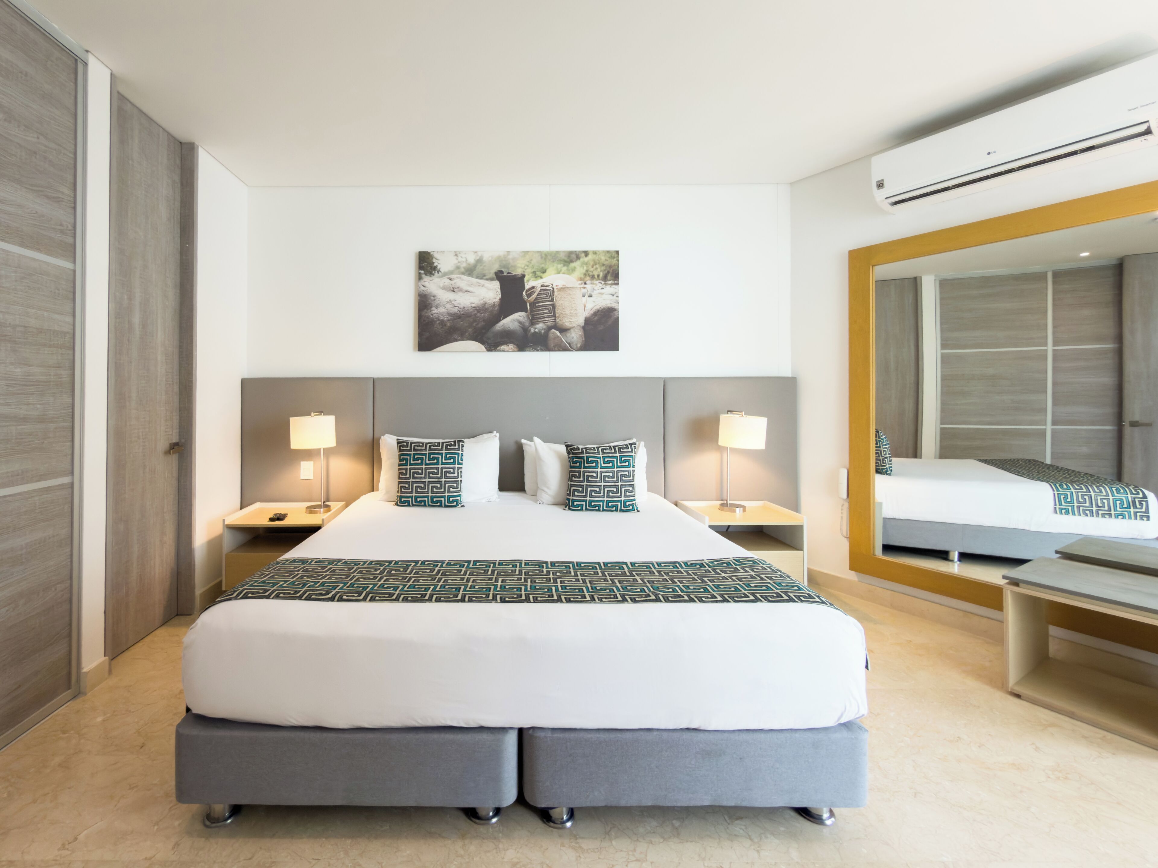 A modern hotel room with a large bed, two bedside tables, a wall-mounted air conditioner, and a large mirror.