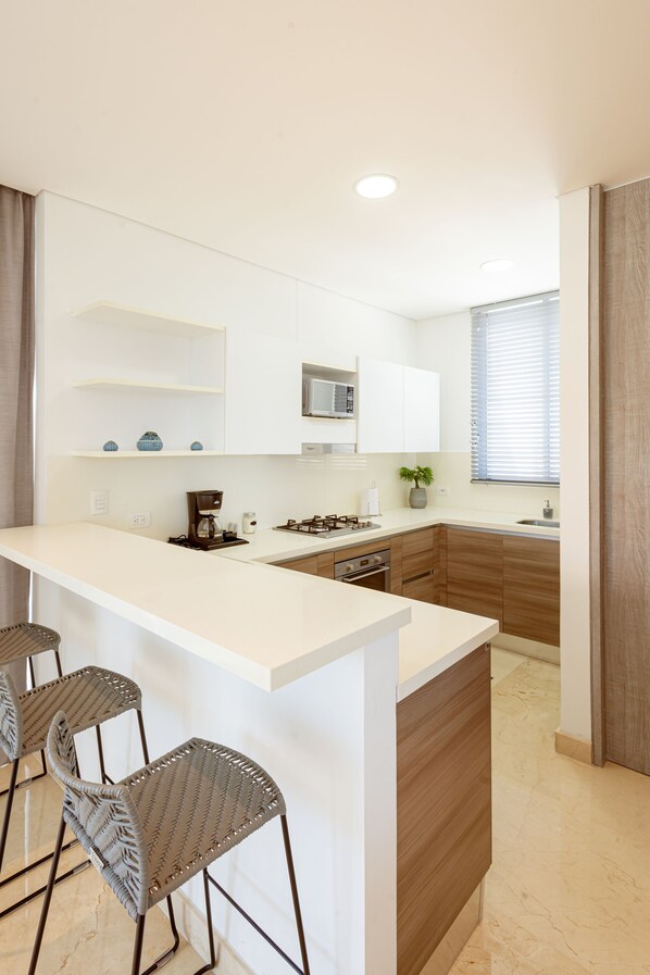 A modern kitchen with a white countertop, wooden cabinets, a microwave, and a coffee maker. There are three metal chairs with a woven design. - Grand Marina Santa Marta (Santa Marta)