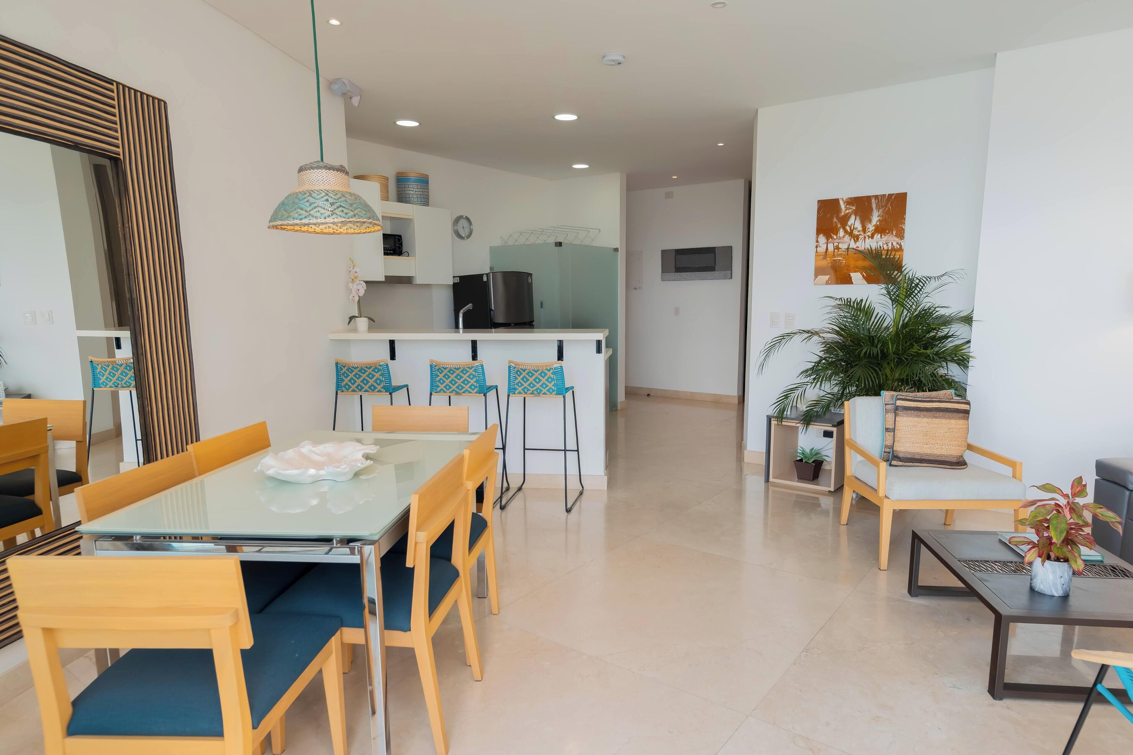 A modern living room with a glass dining table, wooden chairs, a sofa, and a coffee table. There is a kitchen area with a refrigerator, microwave, and a hanging light fixture.
