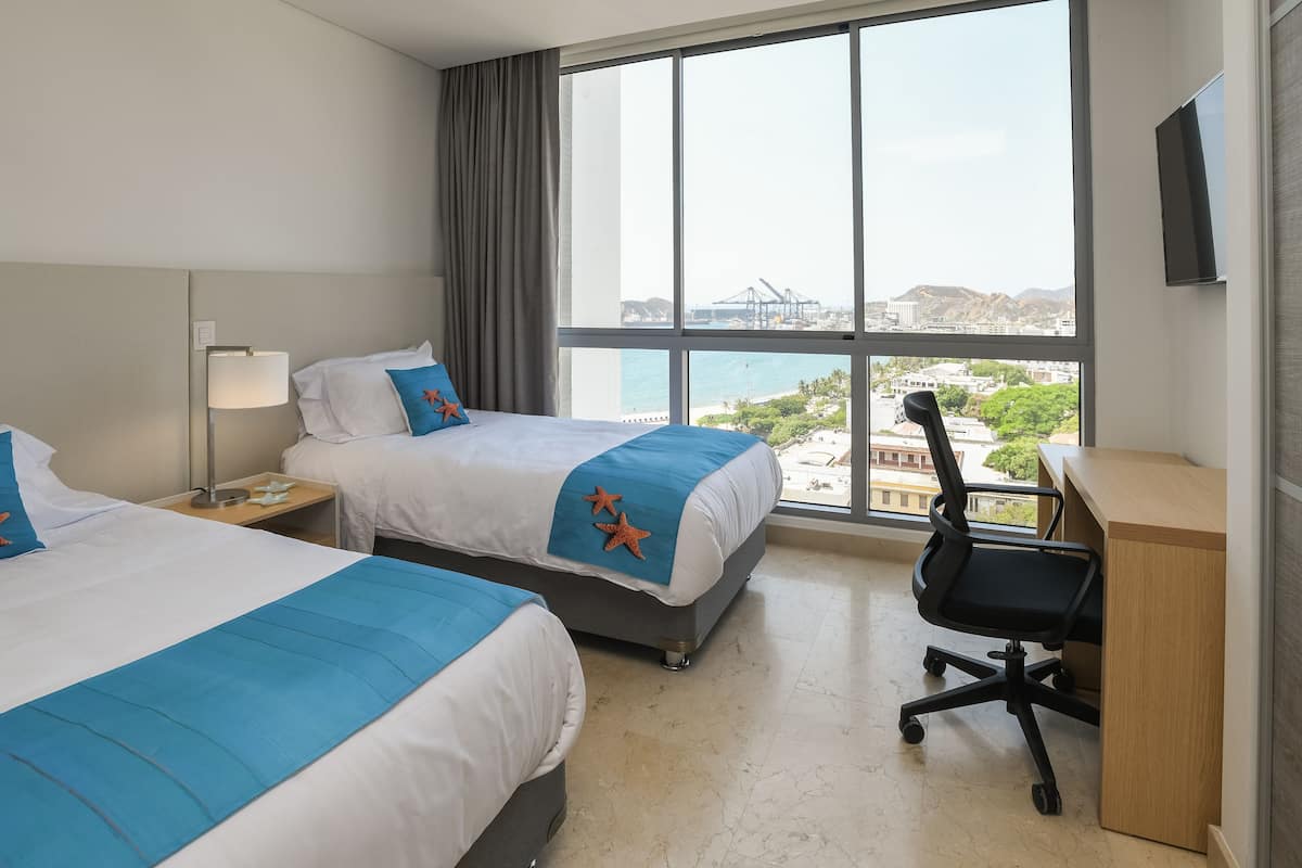 A hotel room with two beds, a desk, a chair, a TV, and a large window with a view of the city and mountains.