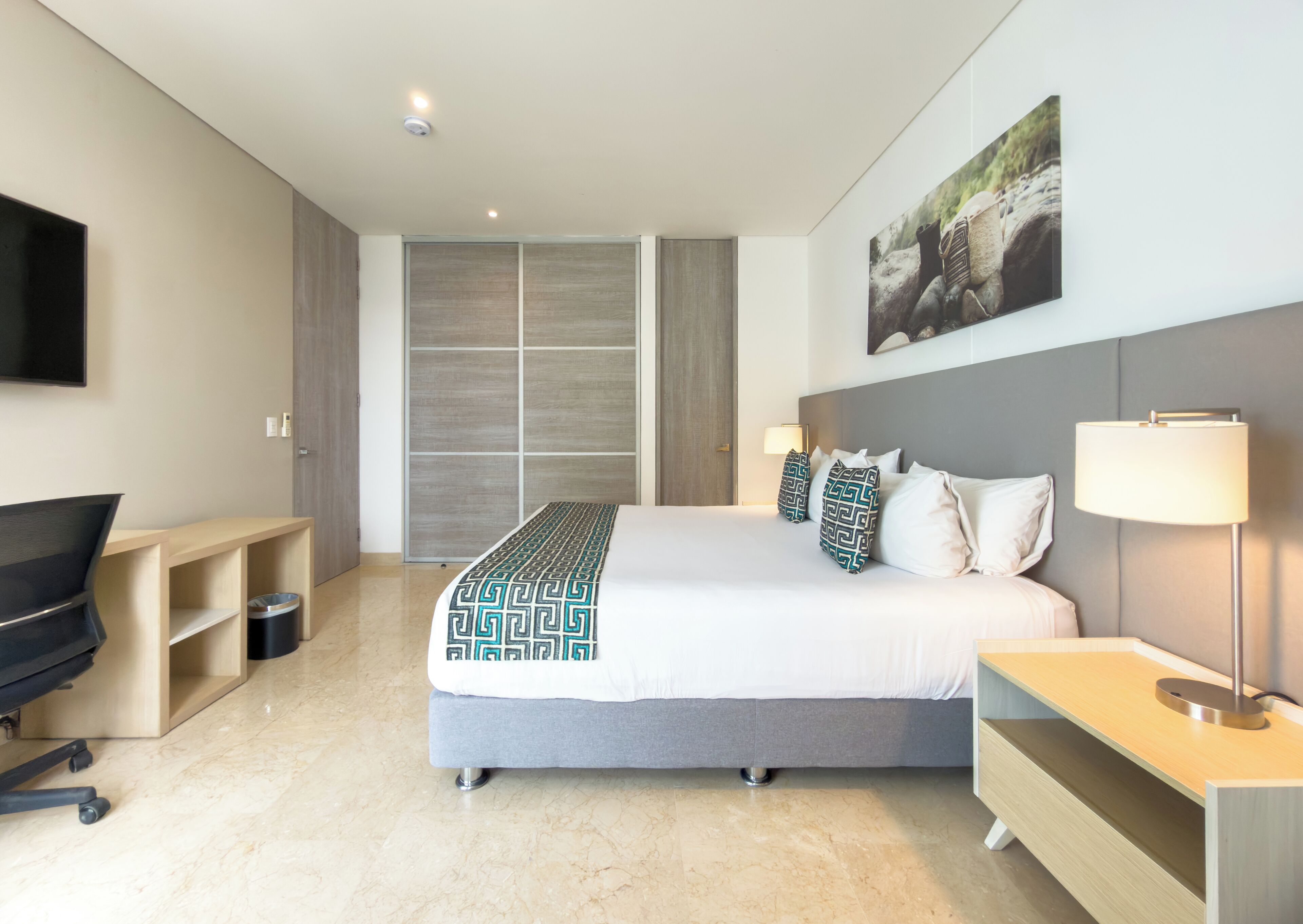 A modern hotel room with a bed, bedside tables, a desk, a TV, and a large window.