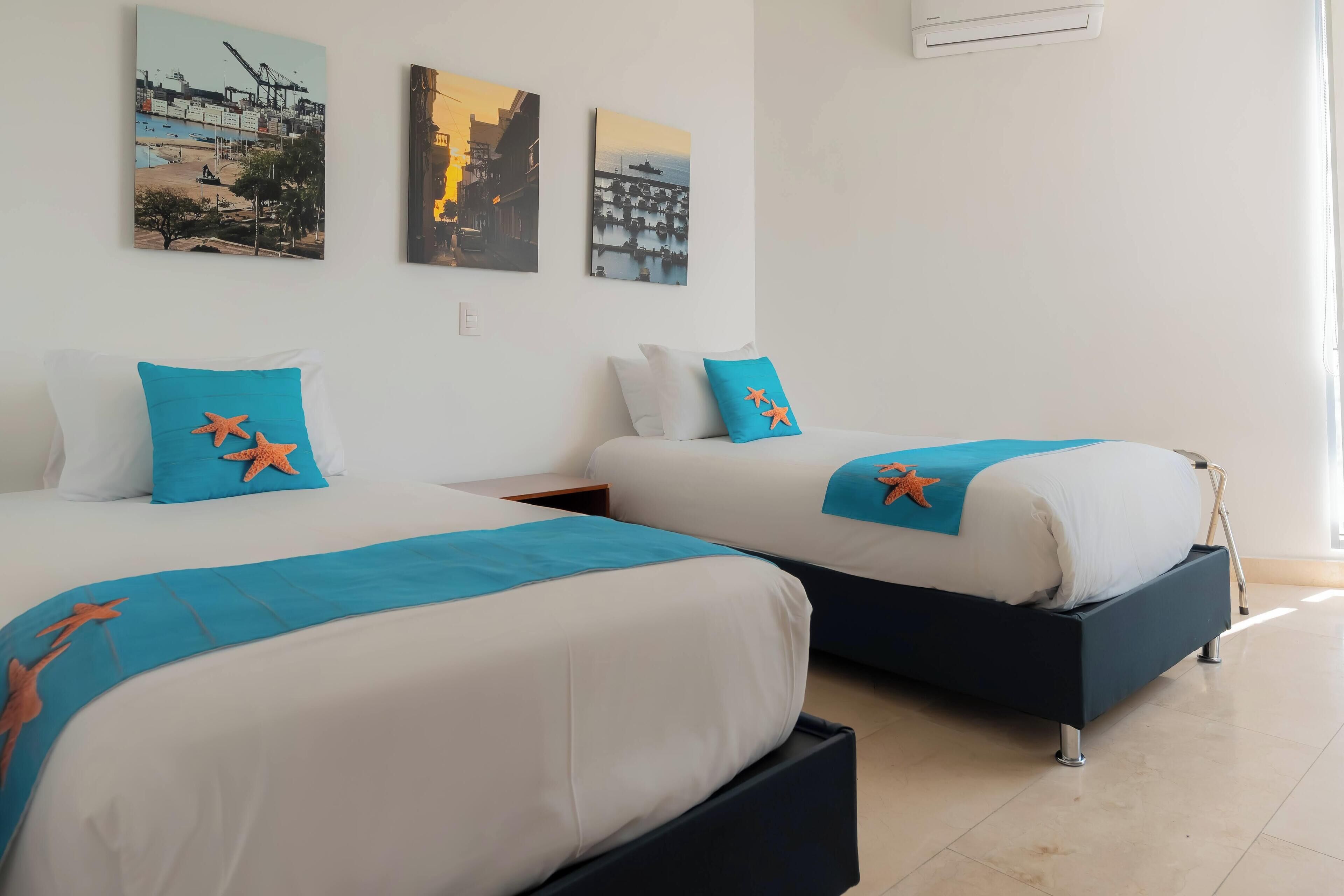 Ein Hotelzimmer mit zwei Betten, jeweils mit blauer Bettdecke und Seesternkissen. An der Wand hängen drei Kunstwerke, die Stadtansichten zeigen.