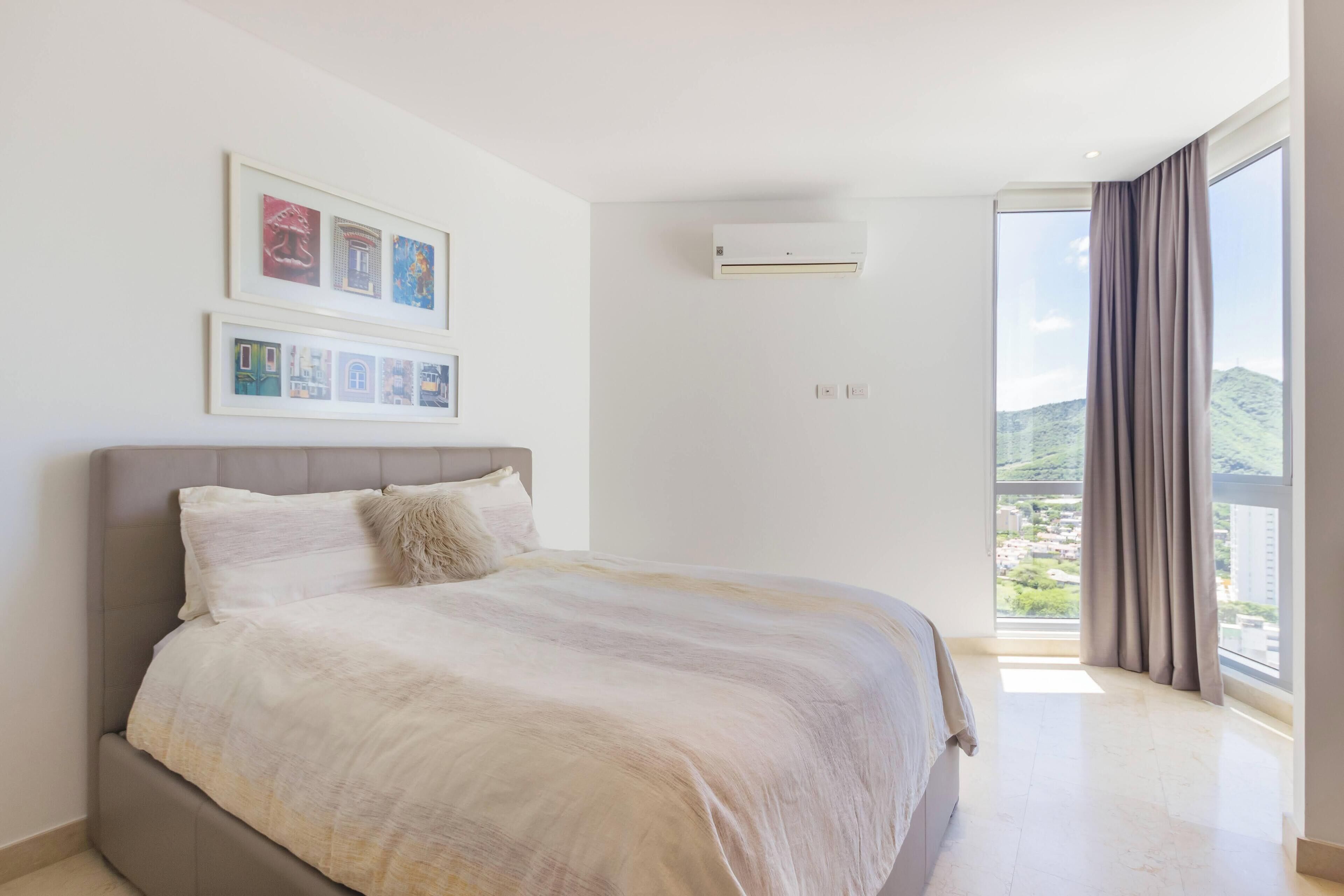 A modern bedroom with a large bed, a mounted air conditioner, and a view of a cityscape through large windows.