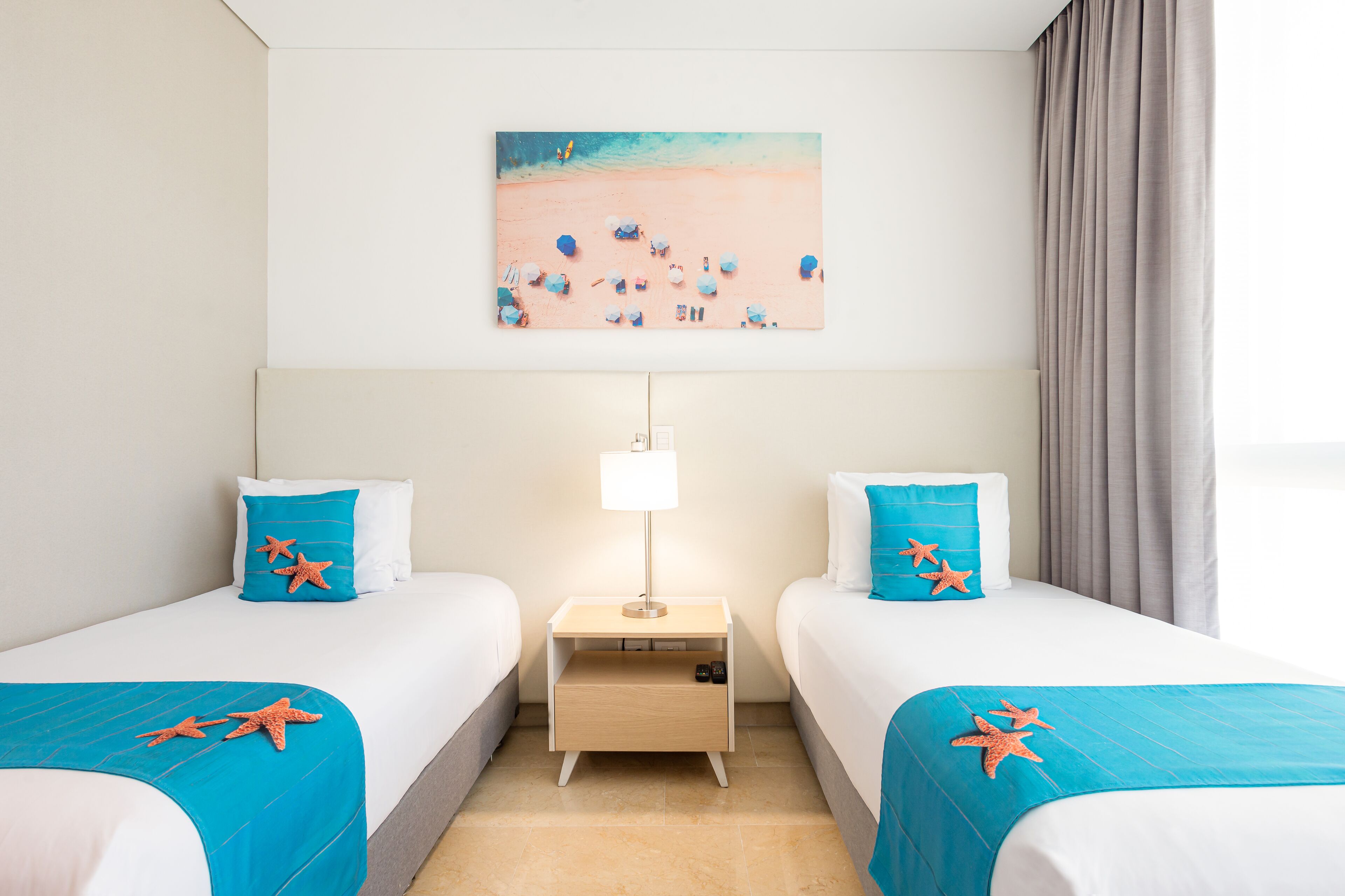 A hotel room with two beds, a nightstand with a lamp, and a beach-themed painting on the wall.