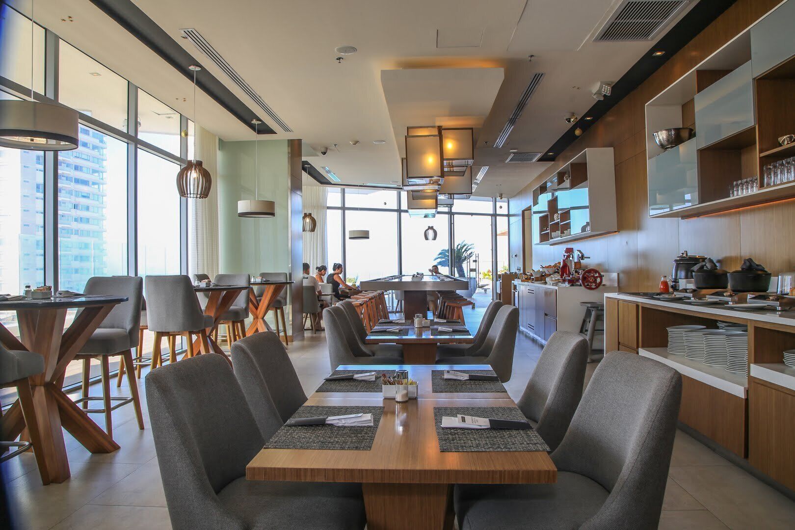 A modern restaurant with a central dining area, a kitchen visible to the right, and large windows providing natural light.