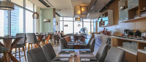 A modern restaurant with a central dining area, a kitchen visible to the right, and large windows providing natural light.