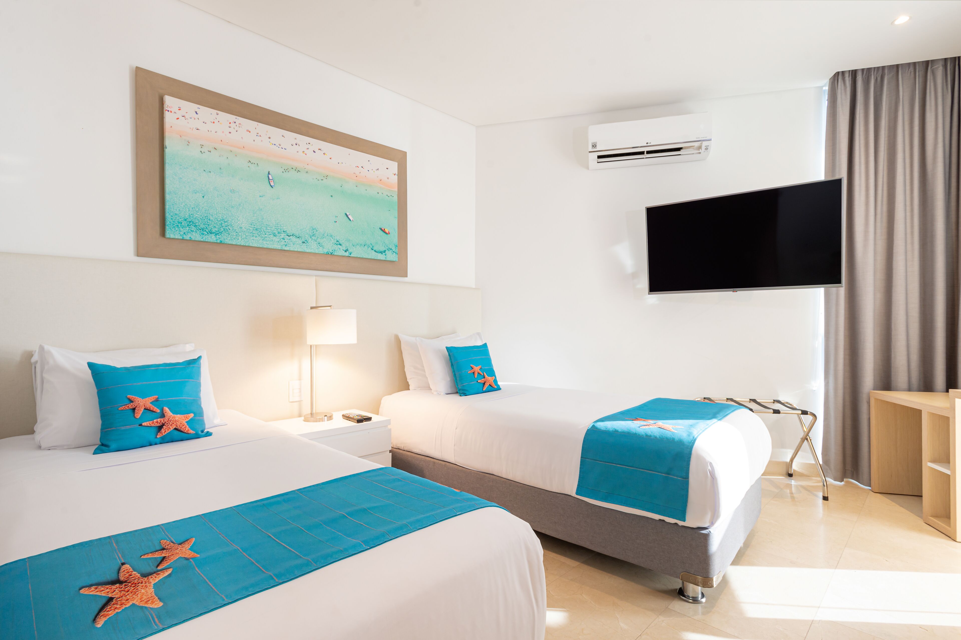 Habitación de hotel con dos camas, televisor, equipo de aire acondicionado y un cuadro con una escena de playa.