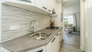 A modern kitchen with a stainless steel sink, a gas stove, and a tiled backsplash. There is a dining area with a sofa and a coffee table visible in the background. - Eureka Park (Renesse)