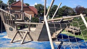 A wooden pirate ship play structure with a rope net, surrounded by a blue and green play area. - Eureka Park (Renesse)