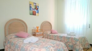 Two single beds with floral bedding, a wicker headboard, a small table with a vase, and a colorful abstract painting on the wall. - Holiday Apartment in Santa Margherita (Santa Margherita Ligure)