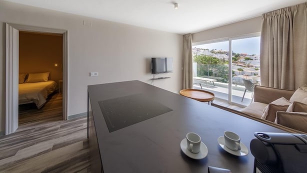 A hotel room with a bed, a TV, a desk with two cups, a chair, and a view of the city. - Costa Adeje Garden (Adeje)