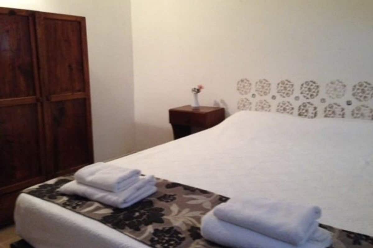 A hotel room with a bed, a wooden wardrobe, a nightstand with a vase, and a patterned wall.