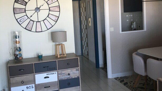 A living room with a large wall clock, a wooden cabinet with drawers, a lamp, and a TV mounted on the wall. - 47m2 duplex apartment - type 2 with terrace (Frejus)