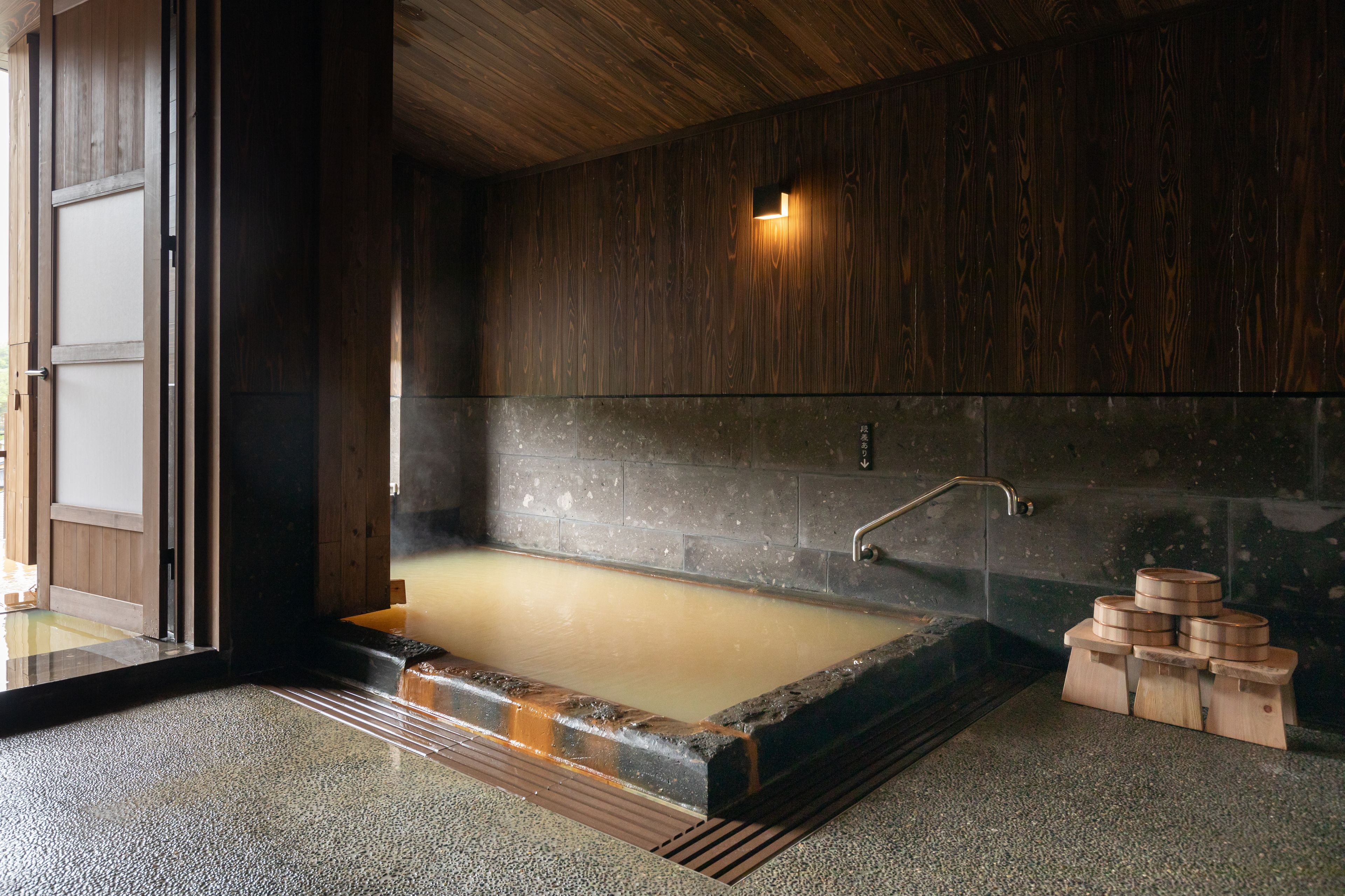 A traditional Japanese onsen with a wooden interior, a stone wall, and a wooden bench with containers.