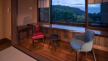 A room with wooden flooring, a red chair, a blue chair, and a stool. There are large windows with a view of a forested area.