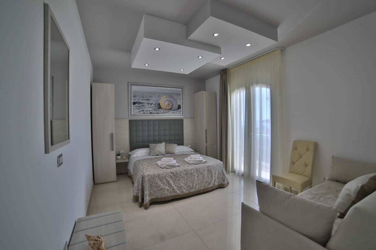A modern bedroom with a large bed, a headboard with built-in lighting, a bedside table, a chair, and a window with curtains.