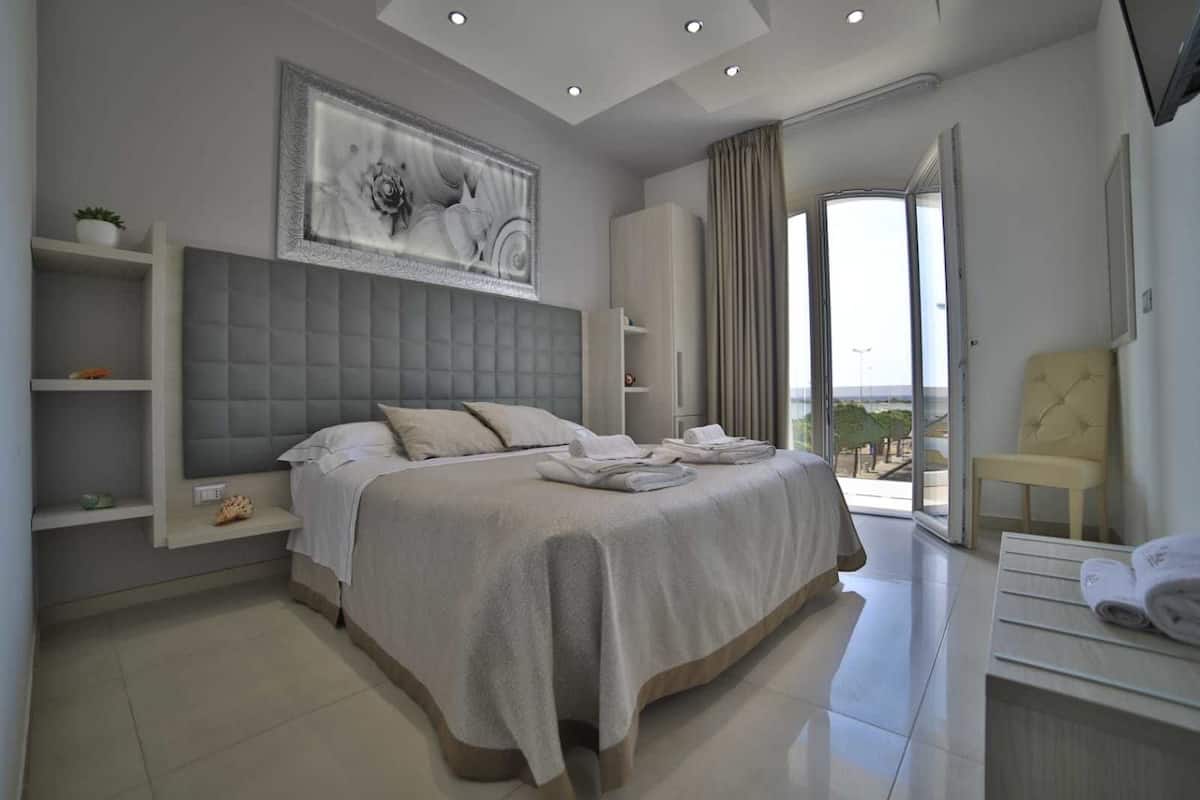 A modern hotel room with a large bed, a built-in wardrobe, a seating area with a chair, and a view of the outdoors.