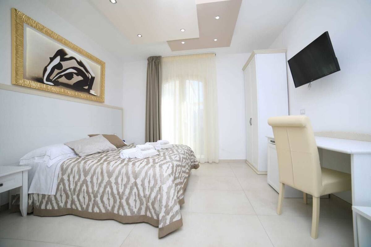 A bedroom with a bed, a desk, a chair, a TV, and a painting on the wall.