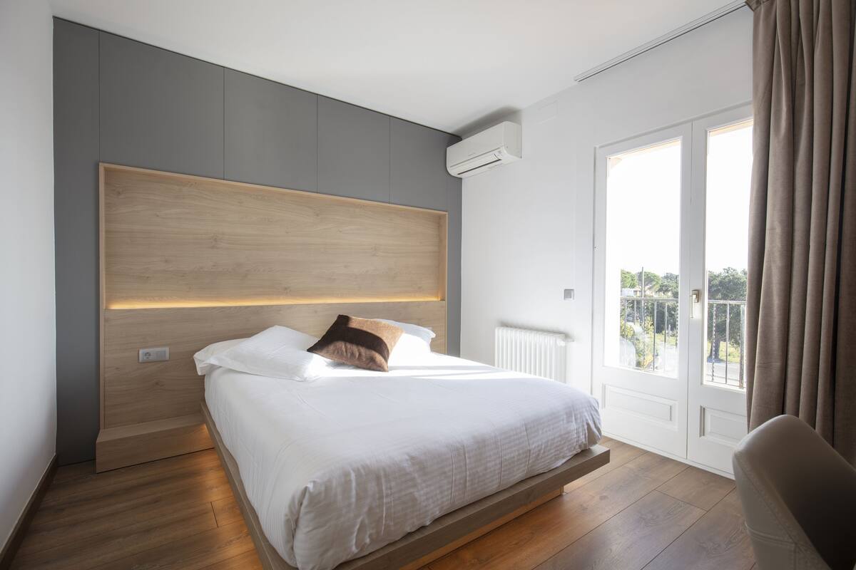 A modern bedroom with a wooden headboard, a large window with curtains, and a view of the outdoors.