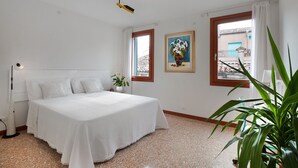 A bedroom with a bed, a nightstand, a potted plant, a painting, and a window with a view of buildings. - Grimaldi Apartments - Ca Sole (Venice)
