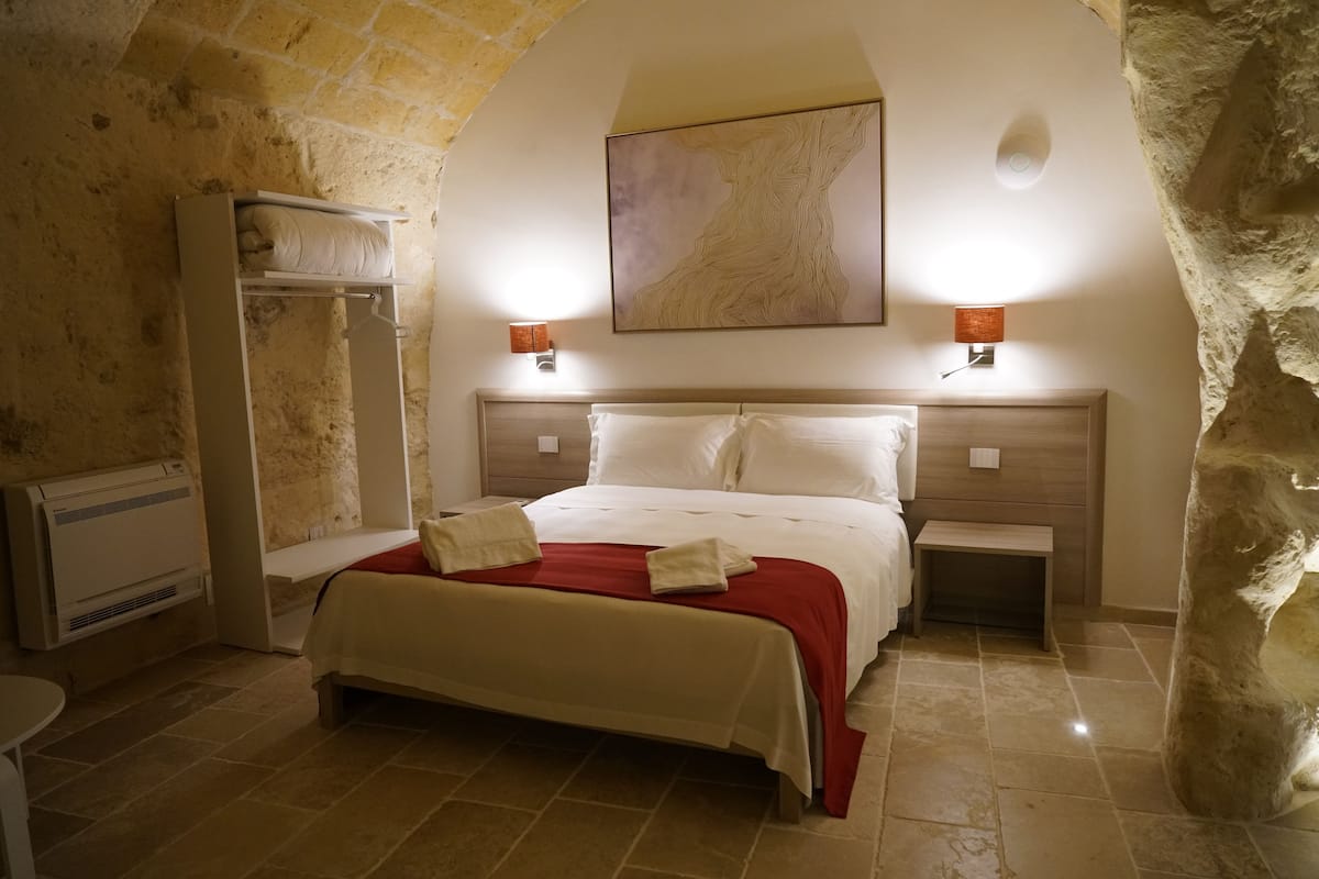 A bedroom with a stone wall, a bed with white linens, two bedside lamps, and a large abstract painting on the wall.