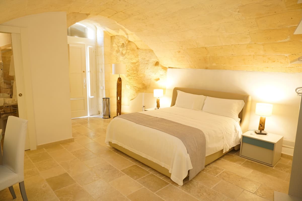 A bedroom with a large bed, a nightstand, a chair, and a stone wall.