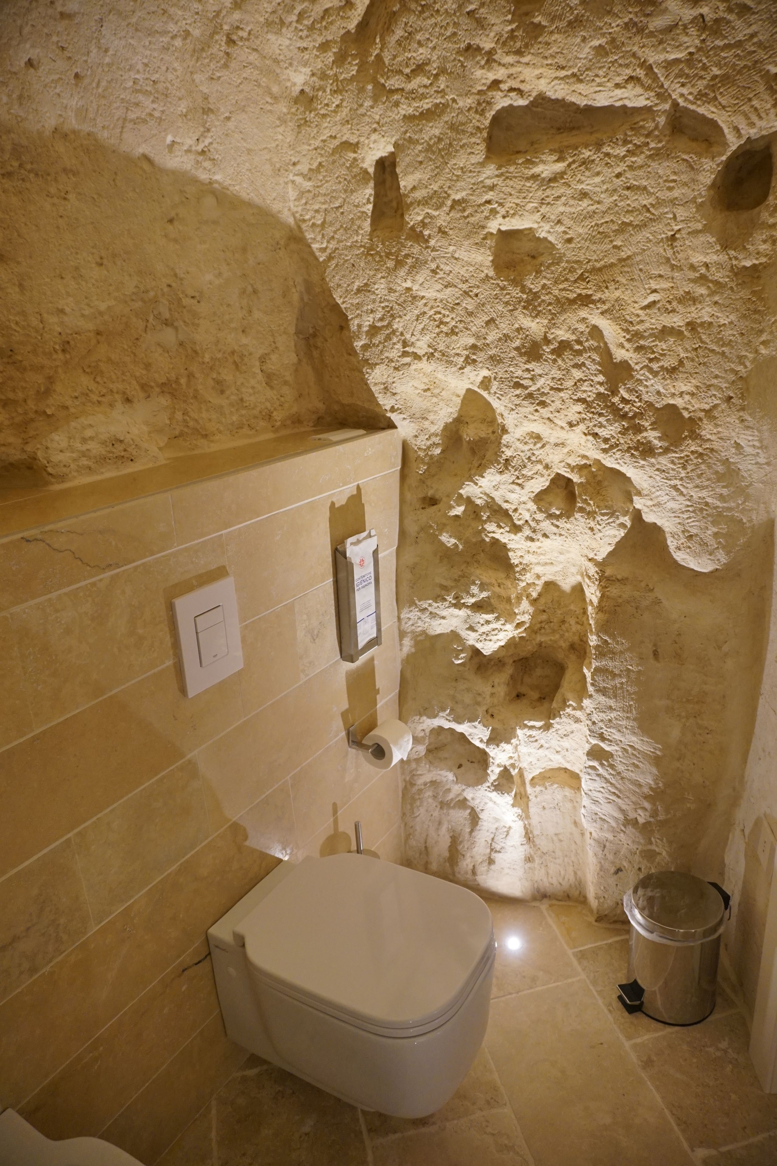 Une salle de bain avec un mur en pierre, des toilettes, une poubelle et un interrupteur fixé au mur.