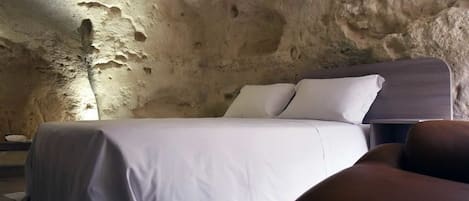 A bedroom with a bed against a textured stone wall, a nightstand, and a brown armchair.