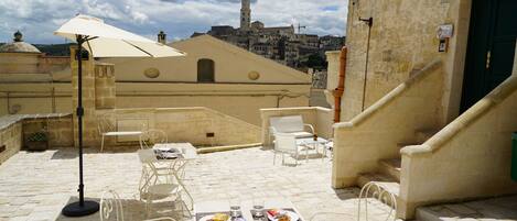 Une terrasse extérieure avec un sol en pierre, des chaises en métal blanc, une table avec des assiettes et des verres, et un grand parasol. Au fond, on aperçoit un bâtiment historique doté d’une tour.