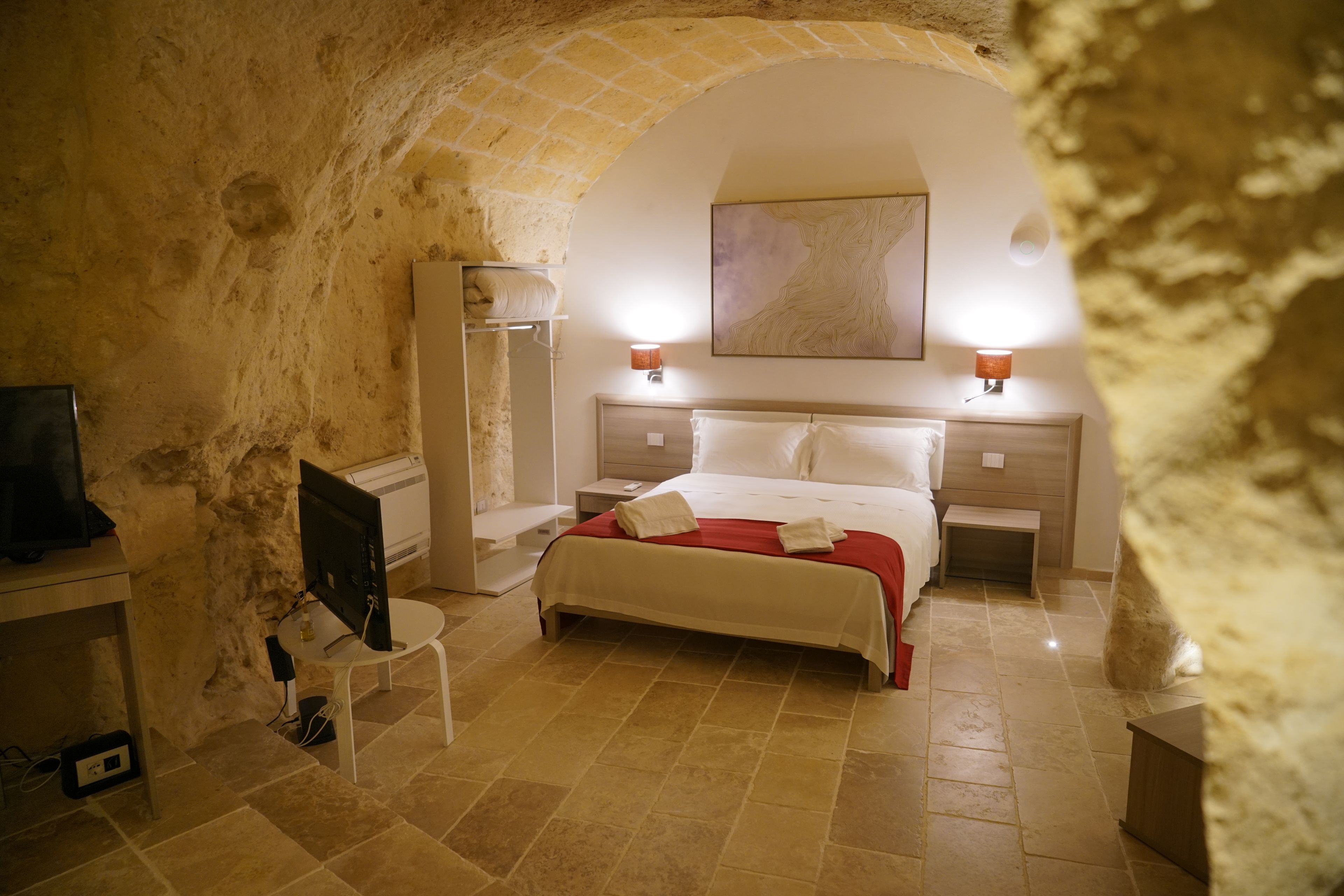 A hotel room with a stone arch, a bed with white linens, a flat-screen TV, a bedside table, and a lamp.