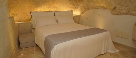 Quarto com parede de pedra, cama com colcha bege, mesa de cabeceira e criado-mudo.