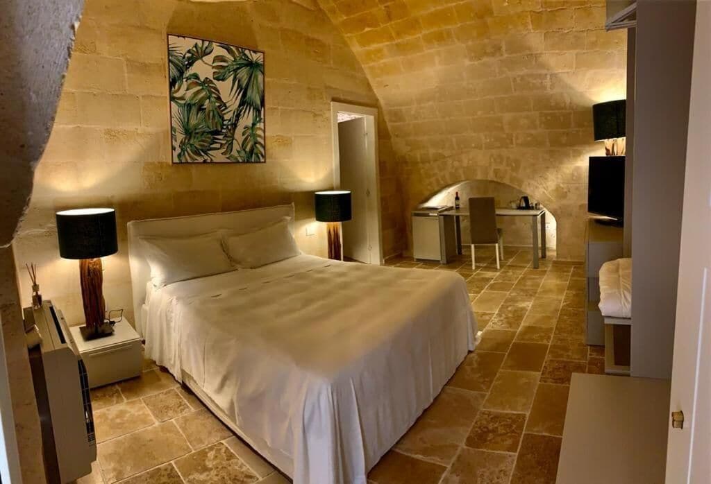 A bedroom with a large bed, two bedside tables with lamps, a small desk, and a chair. The room has stone walls and a sloped ceiling.