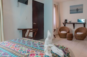 Un dormitorio con una cama, un escritorio, dos sillas de mimbre, una mesita con cafetera y un cuadro en la pared.